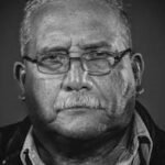 Black and white portrait of a senior man wearing glasses and a jacket.